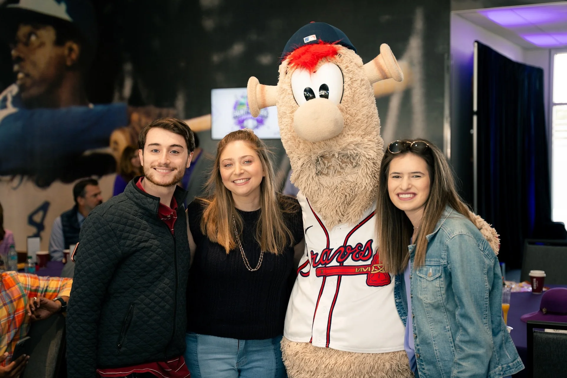 Three young adults (two women and one man) posing with the Atlanta Braves mascot, a large anthropomorphic bear wearing a baseball cap and jersey, indoors at a social event.