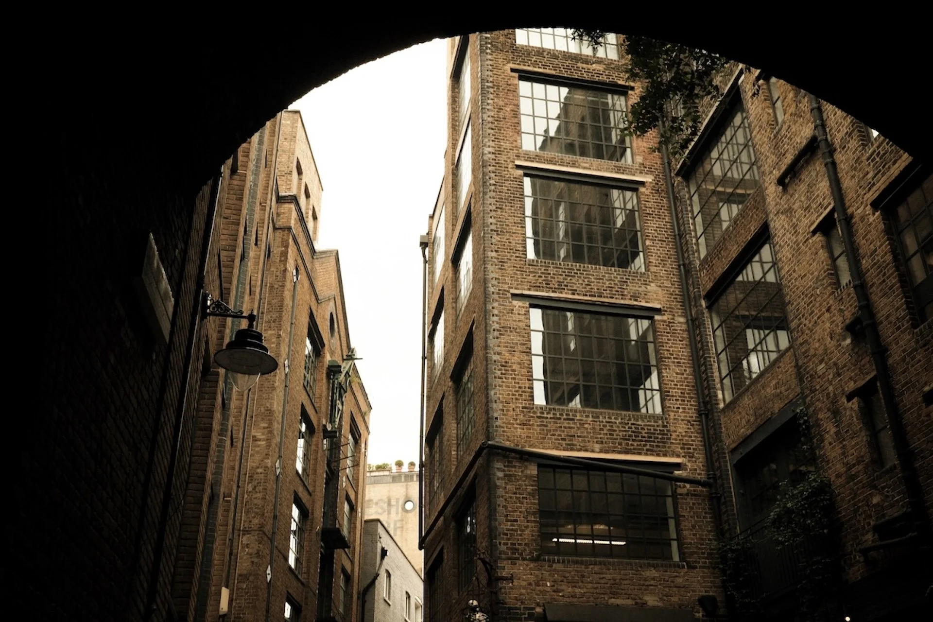 View through an archway showing brick buildings with large windows on a cloudy day.