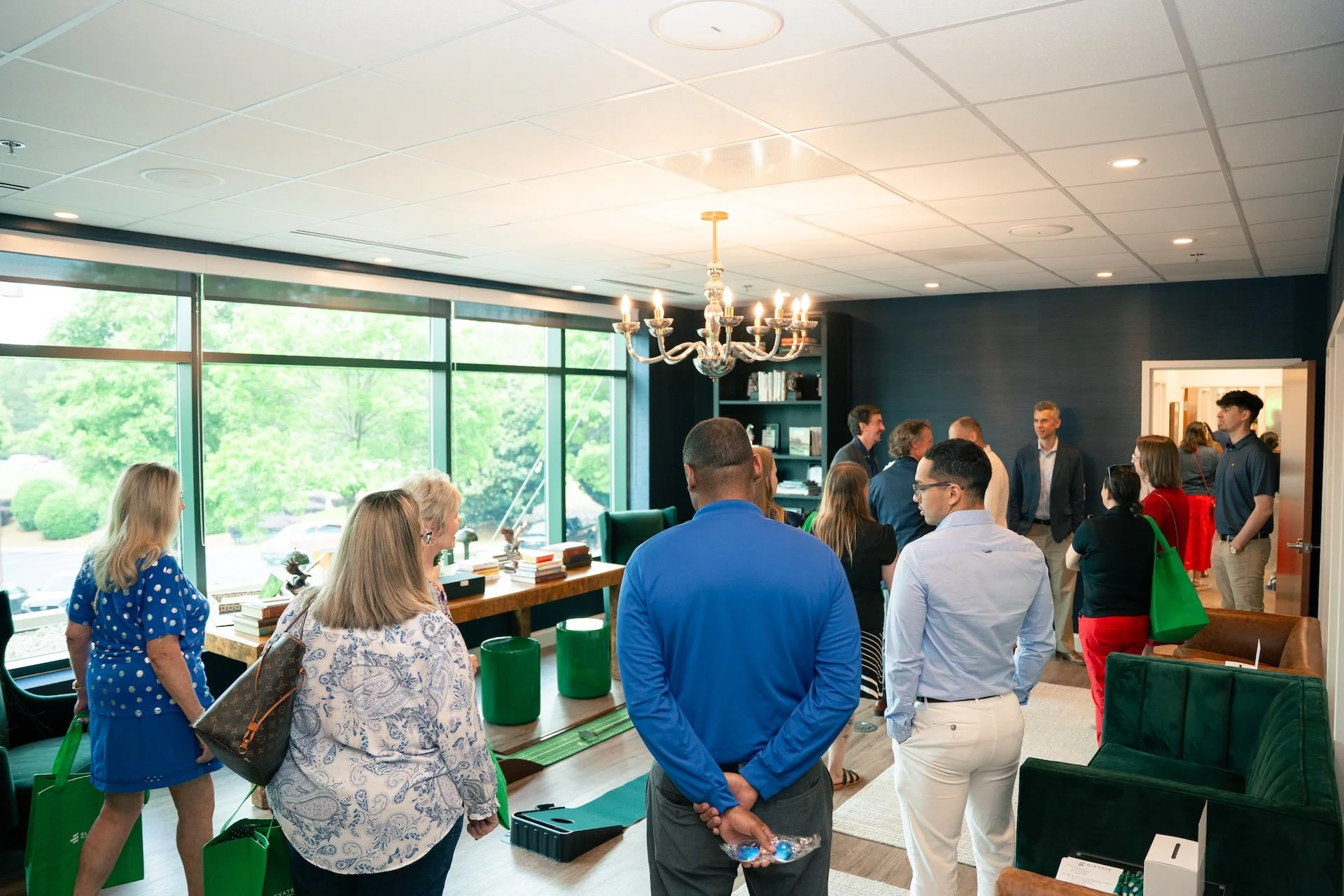 Group of people gathered in a room with large windows, dark walls, and a chandelier, engaging in conversations.