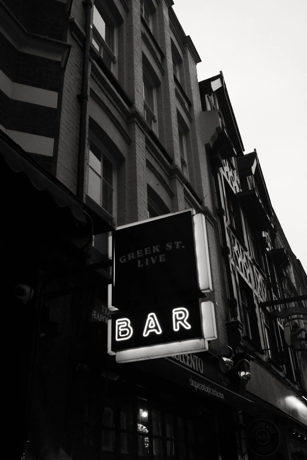 A black and white photo of a city street with a neon sign reading 'BAR' hanging outside a building. The building has brick walls and multiple windows with balconies. There is a street sign that says 'GREEK ST.'.