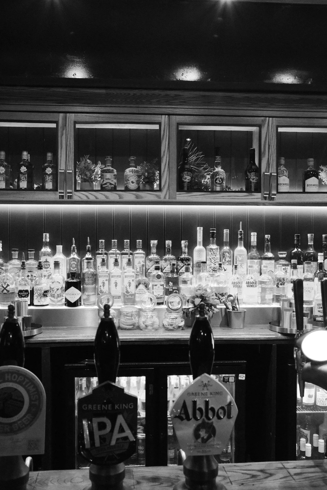 Black and white photo of a bar with various bottles of alcohol on shelves, including gin, vodka, and whiskey, with beer tap handles labeled GBP IPA and Abbot in the foreground.