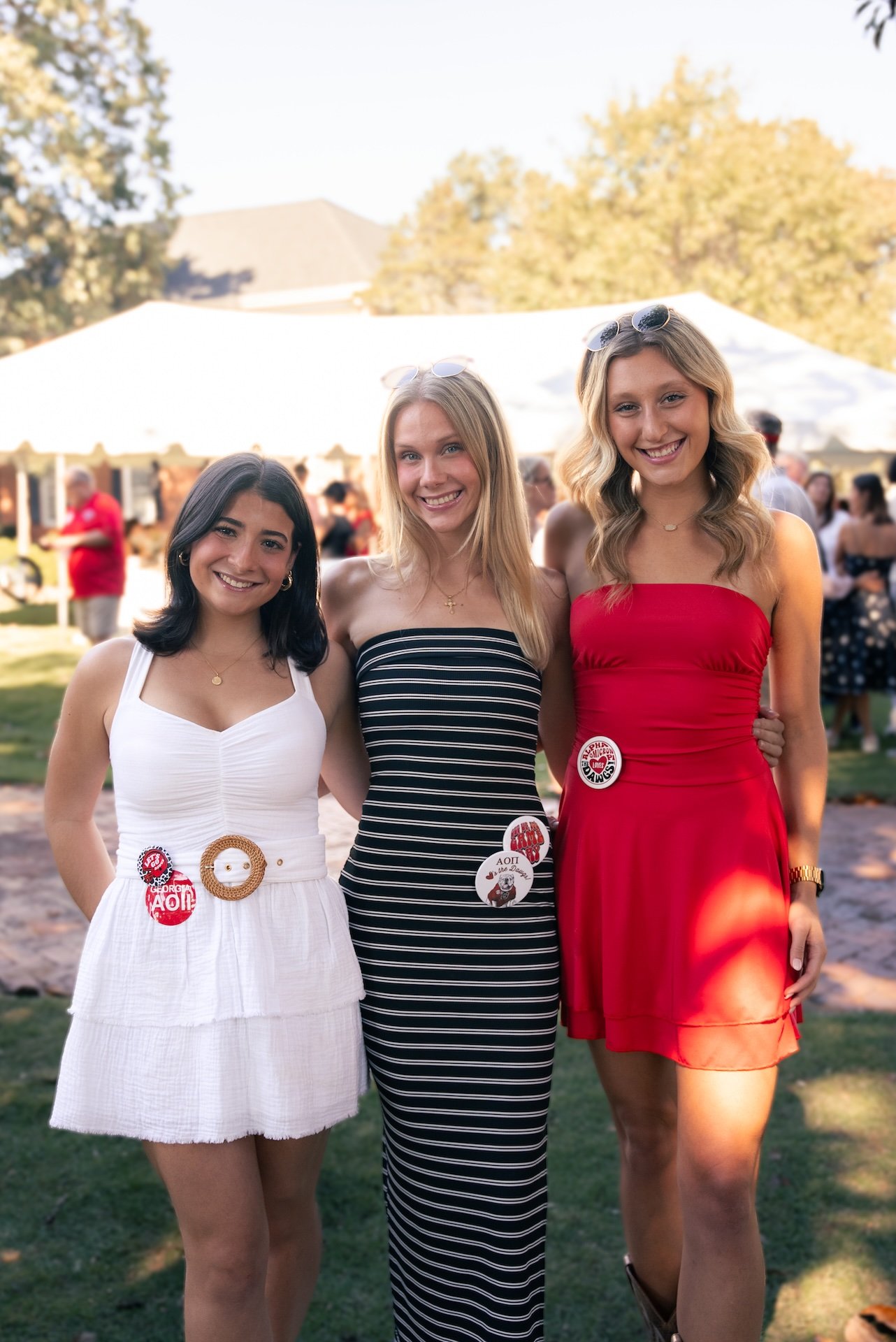 Three young women standing outdoors at a social gathering, smiling at the camera, with a white tent and other people in the background.
