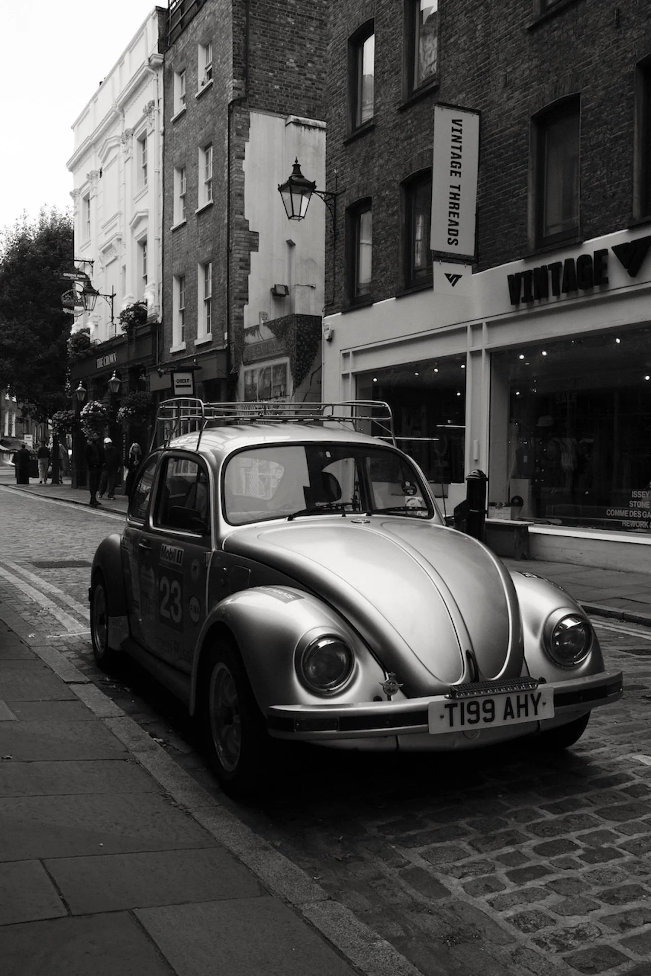 A vintage Volkswagen Beetle parked on a cobblestone street in an urban area, with stores and pedestrians in the background.
