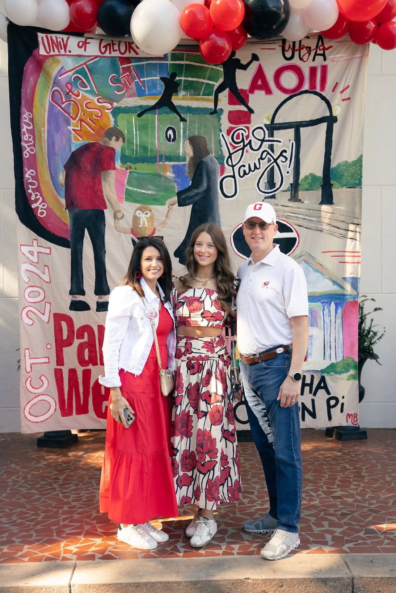 Three people standing in front of a colorful mural at the University of Georgia. The mural features silhouettes of children playing, a rainbow, and the text 'Beautify Miss St!' and 'Go Dawgs!'. The group includes two women and a man, all smiling at t
