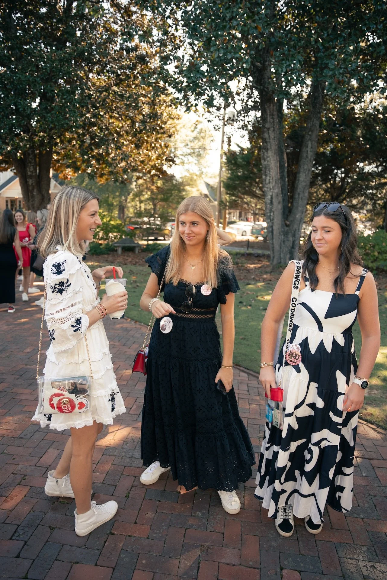 Three young women talking outdoors on a sunny day, dressed in casual summer outfits, with trees and other people in the background.