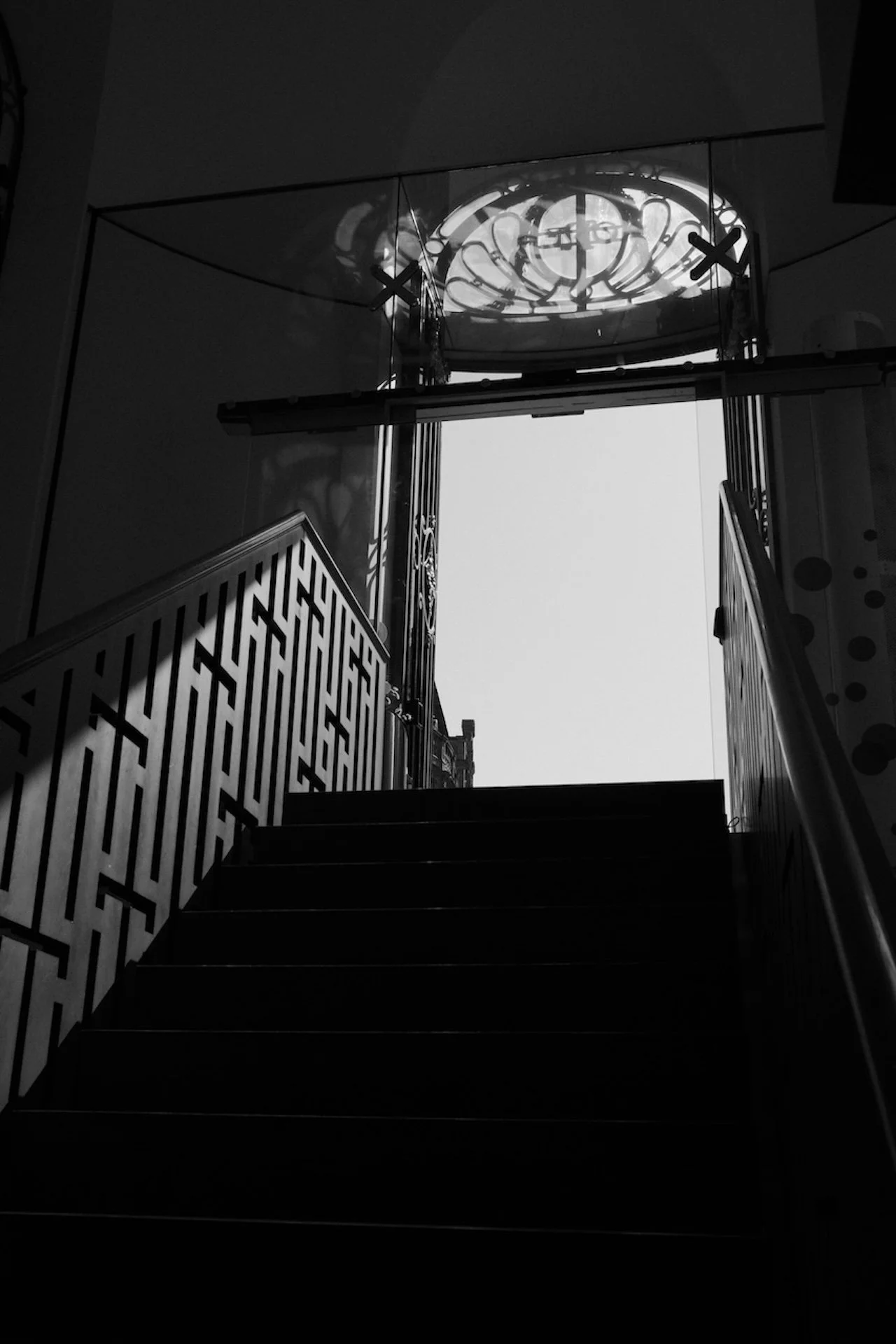 Black-and-white photo of a staircase leading up to an open door with decorative stained-glass window above, sunlight outside casting shadows.