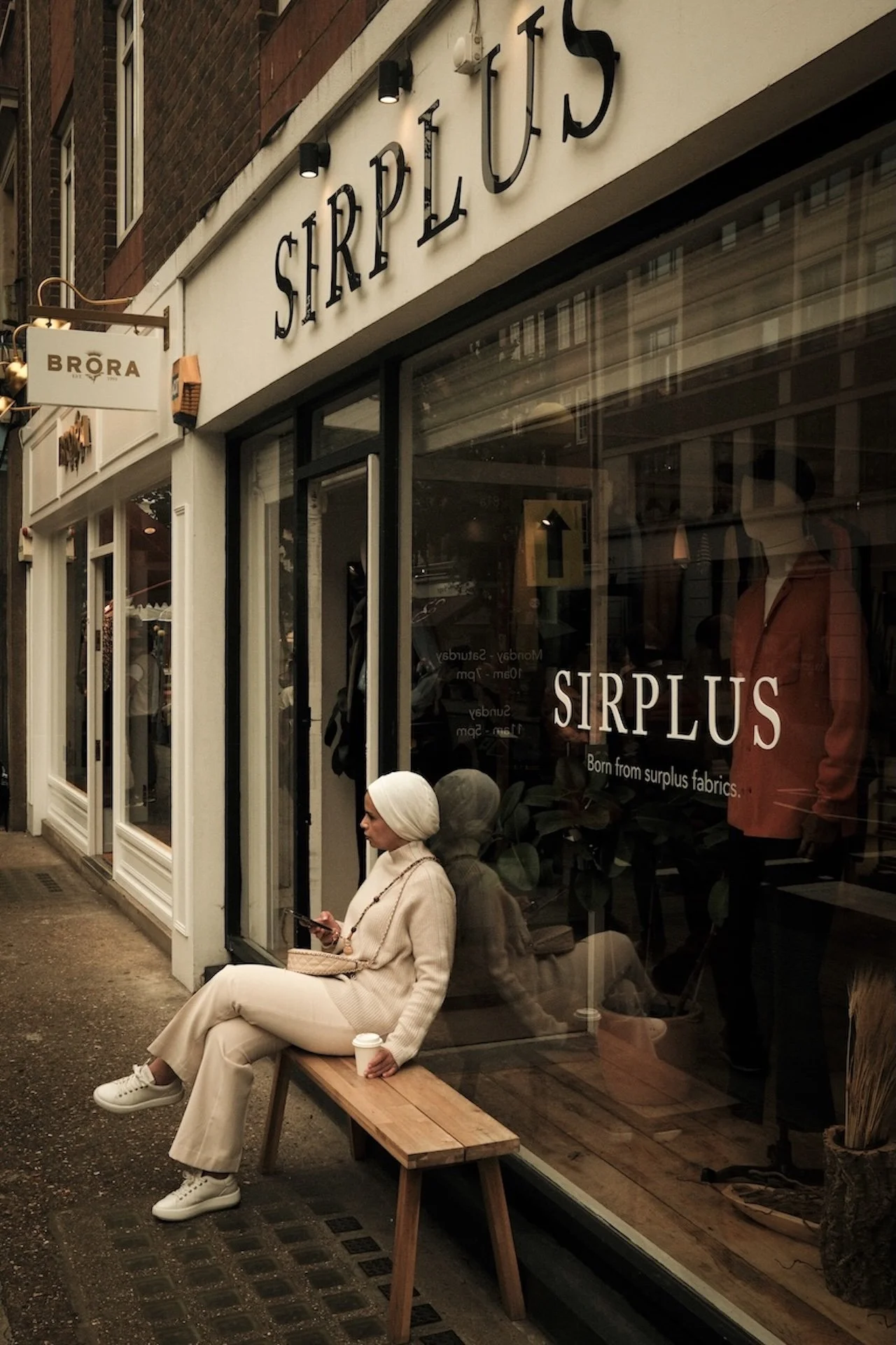 A woman dressed in beige sitting on a wooden bench outside a fashion store called SIRPLUS, looking at her phone, with a coffee cup in her hand.