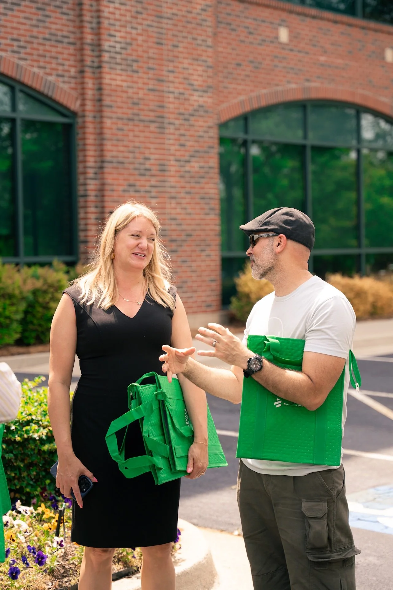 A man and a woman conversing outside near a brick building, holding green reusable bags, with parking lot and green shrubs in the background.