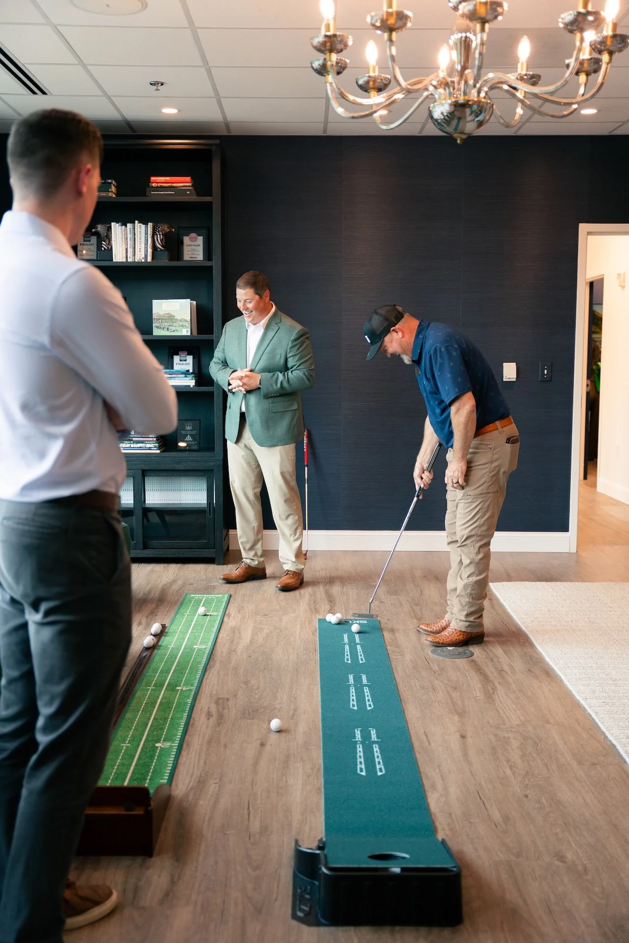 Three men are engaging in a miniature golf game indoors, with two of them preparing to putt and one watching, all standing on wooden floors under a chandelier.