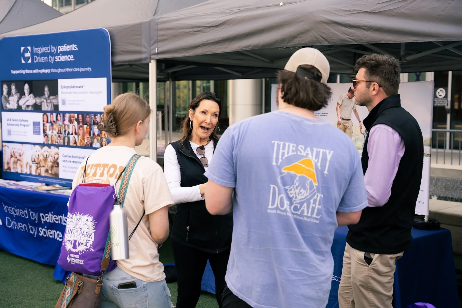 Group of four people talking at an outdoor booth related to epilepsy awareness, with a woman explaining something to a young woman, a man, and another woman, in front of informational posters and a banner.