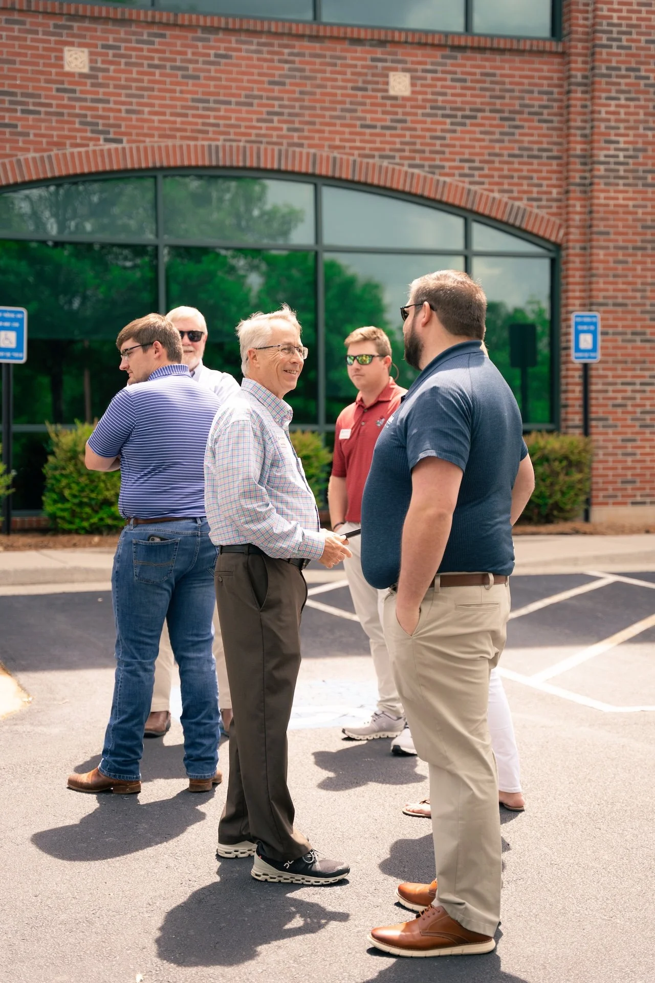 Group of five men standing outside in conversation near a brick building with large windows.