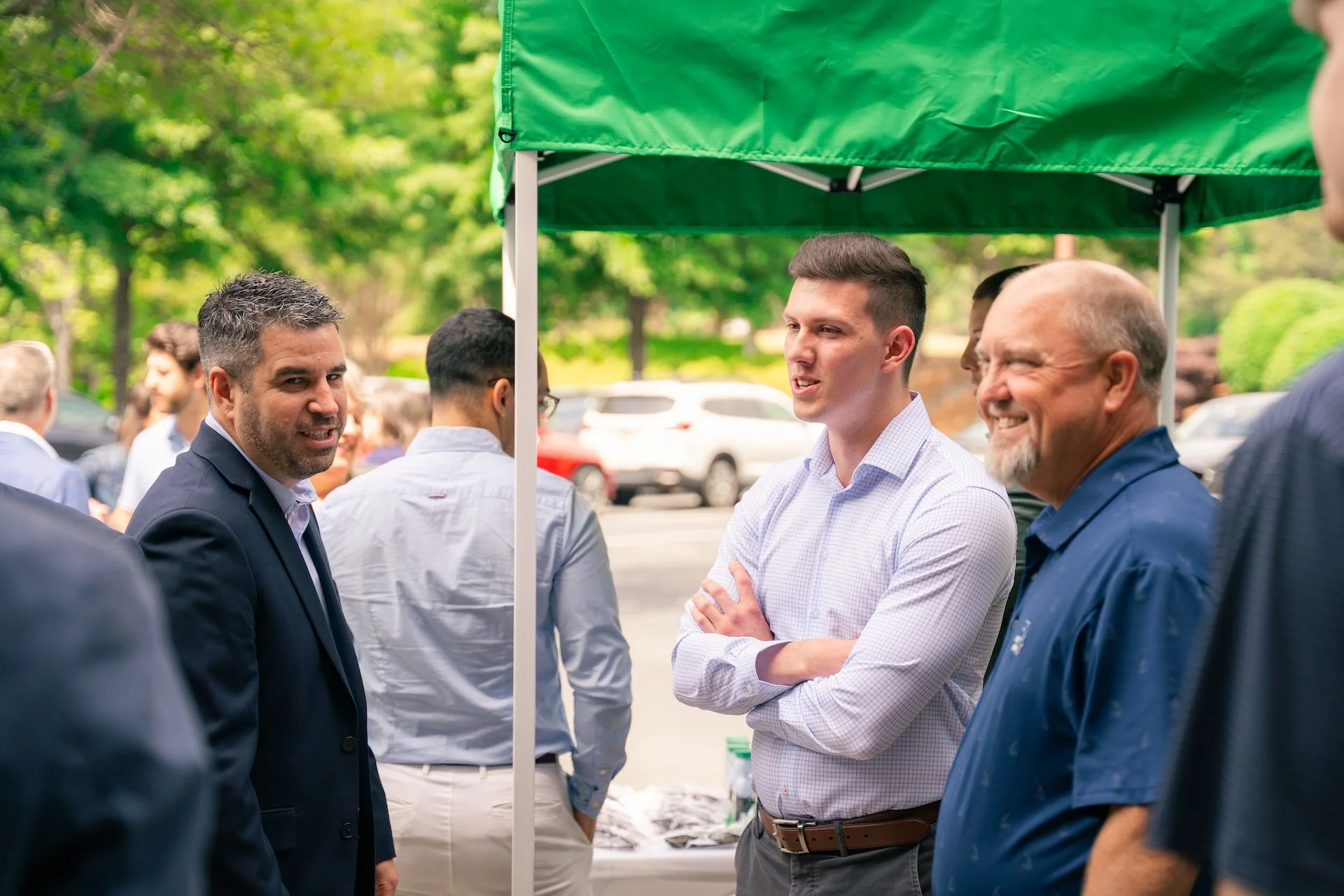 Group of men engaged in conversation at an outdoor event or gathering under a green canopy, with trees and parked cars in the background.