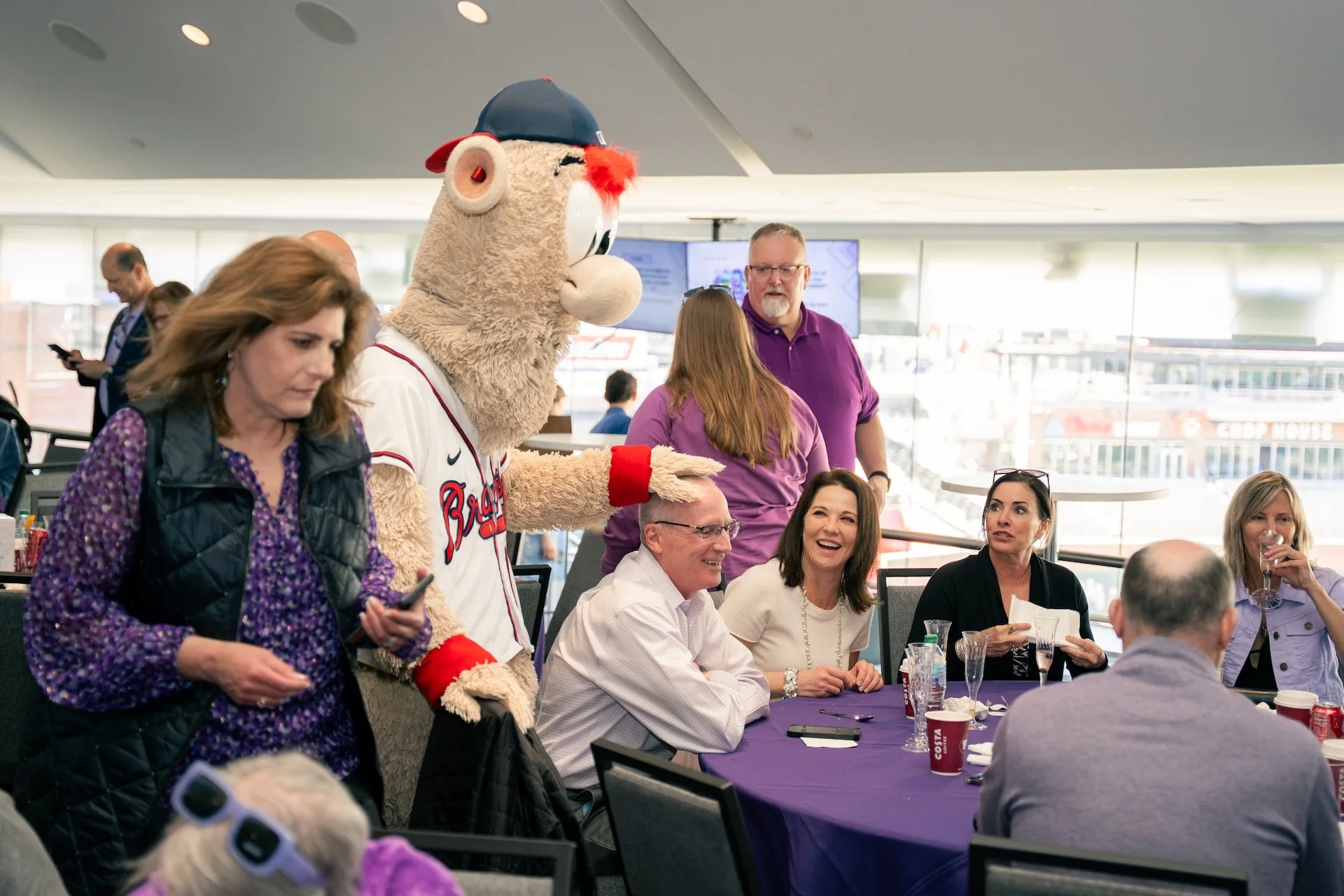 People gathered at a table with a mascot in a room with large windows, some smiling and some engaging with their phones.