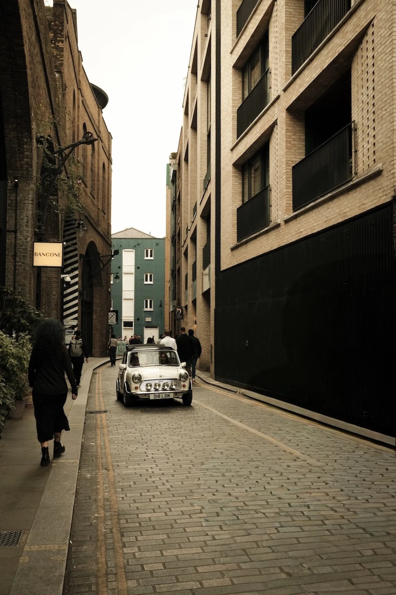 A narrow alleyway in an urban area with brick and modern buildings on both sides. A vintage white car is parked on the cobblestone street, with a few pedestrians walking along the sidewalk.