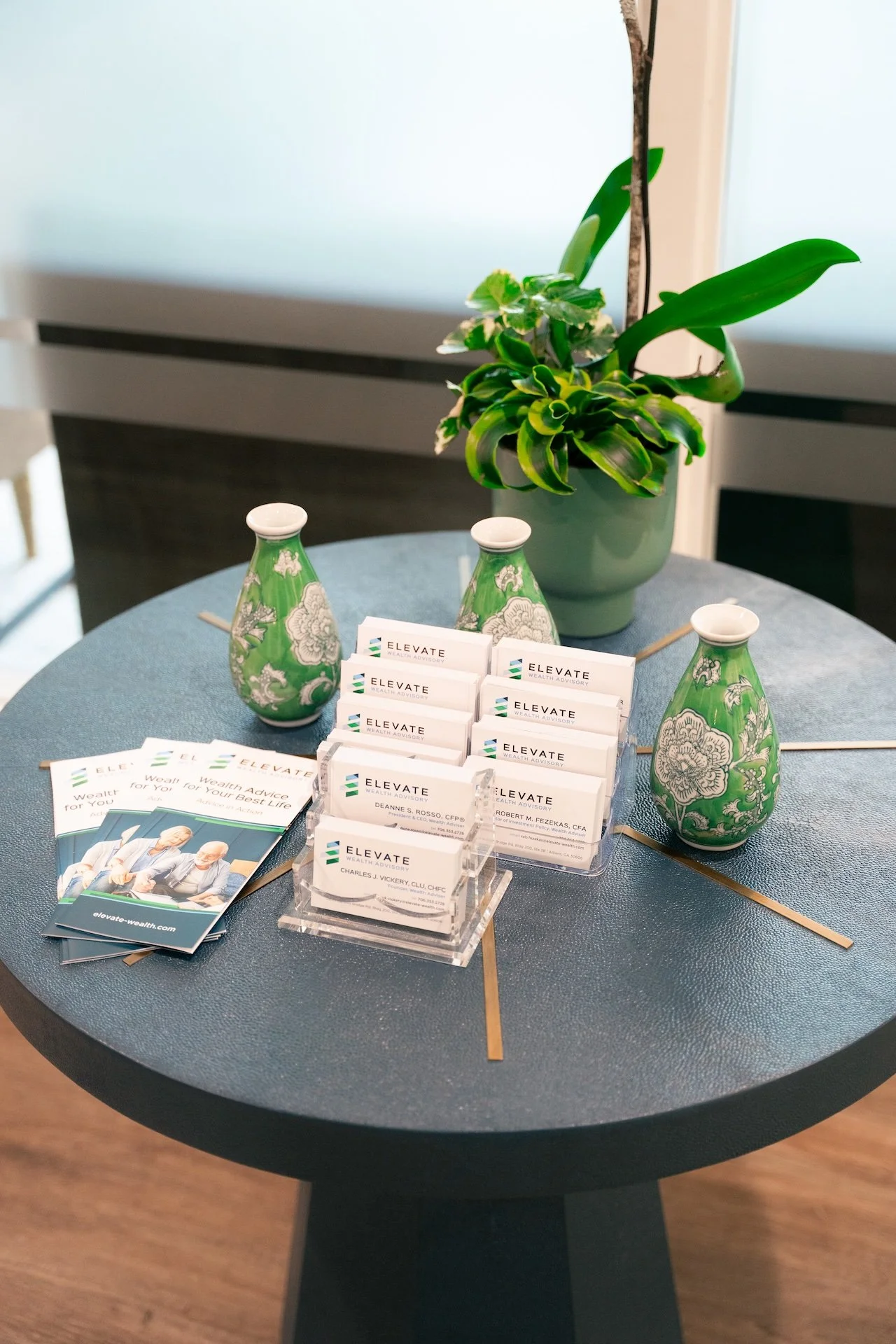 A round table with business cards and pamphlets for Elevate Wealth Advisory, two green decorative vases with white floral patterns, and a potted plant with variegated green leaves.