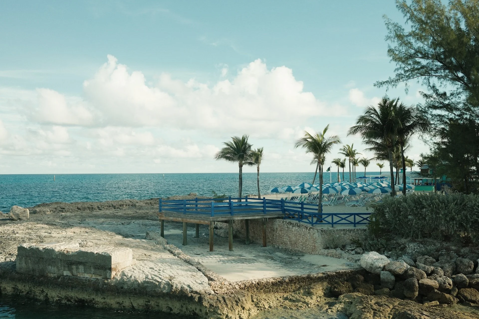 A tropical beach scene with palm trees, blue-striped umbrellas, lounge chairs, rocky shoreline, and the ocean in the background under a partly cloudy sky.