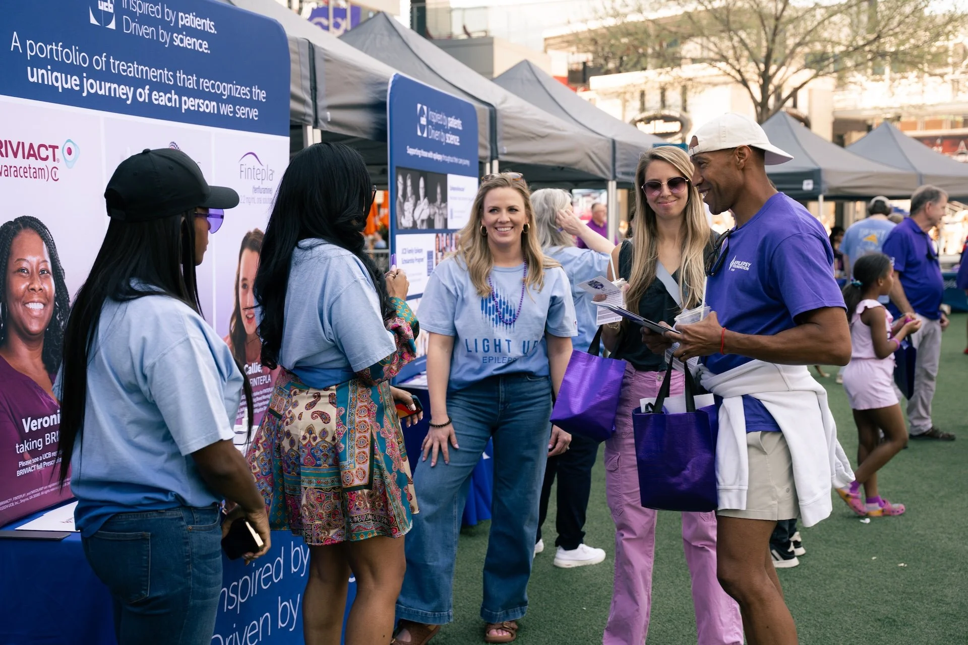 Group of women and a man talking at an outdoor event with tents and informational booths, some wearing matching light blue T-shirts, and one girl in a pink dress in the background.