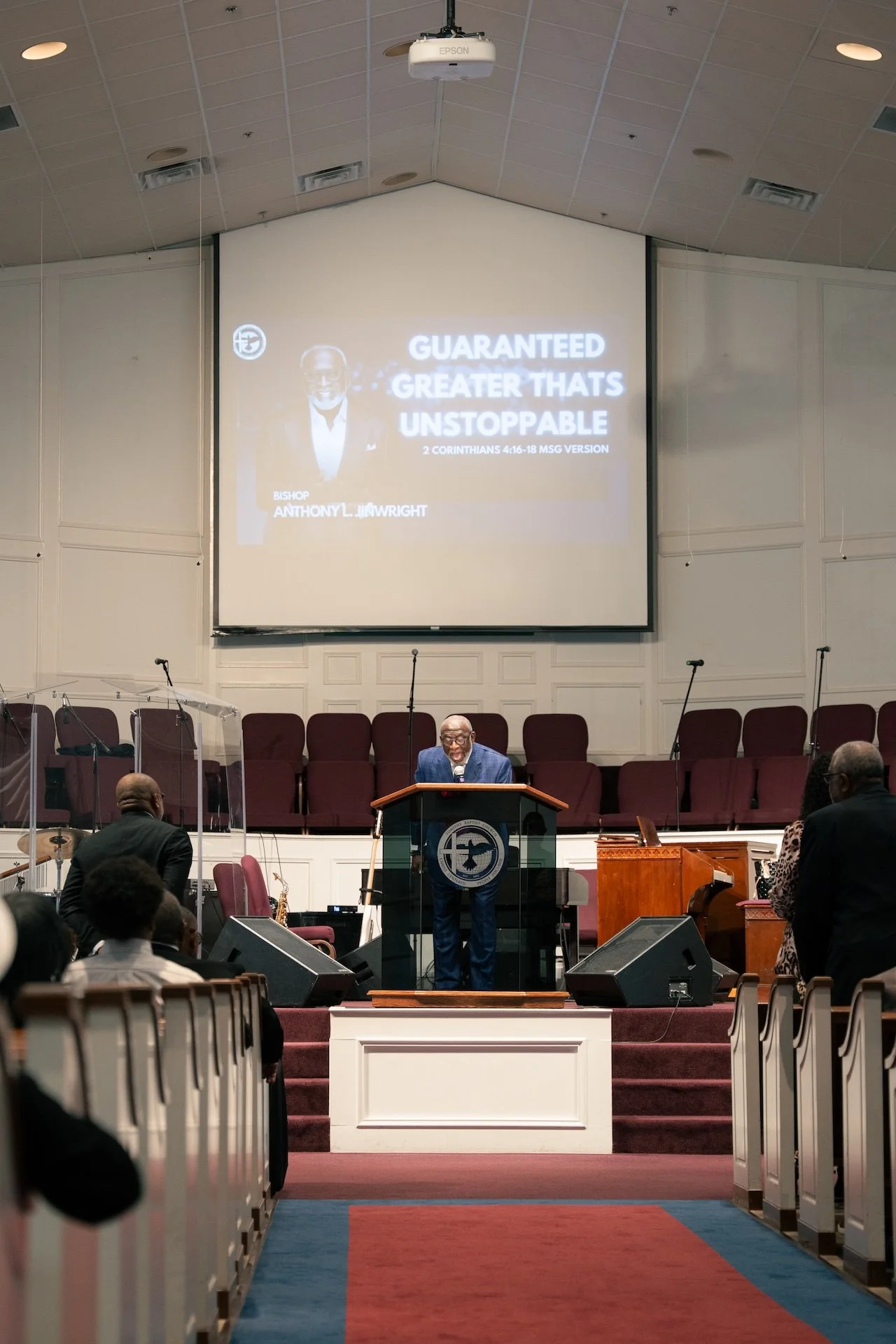 A church service with a preacher standing at the pulpit in front of a large screen displaying a sermon title about 2 Corinthians 4:16-18. The preacher is wearing glasses and a suit. The church interior has a stage with a choir area, musical instrumen