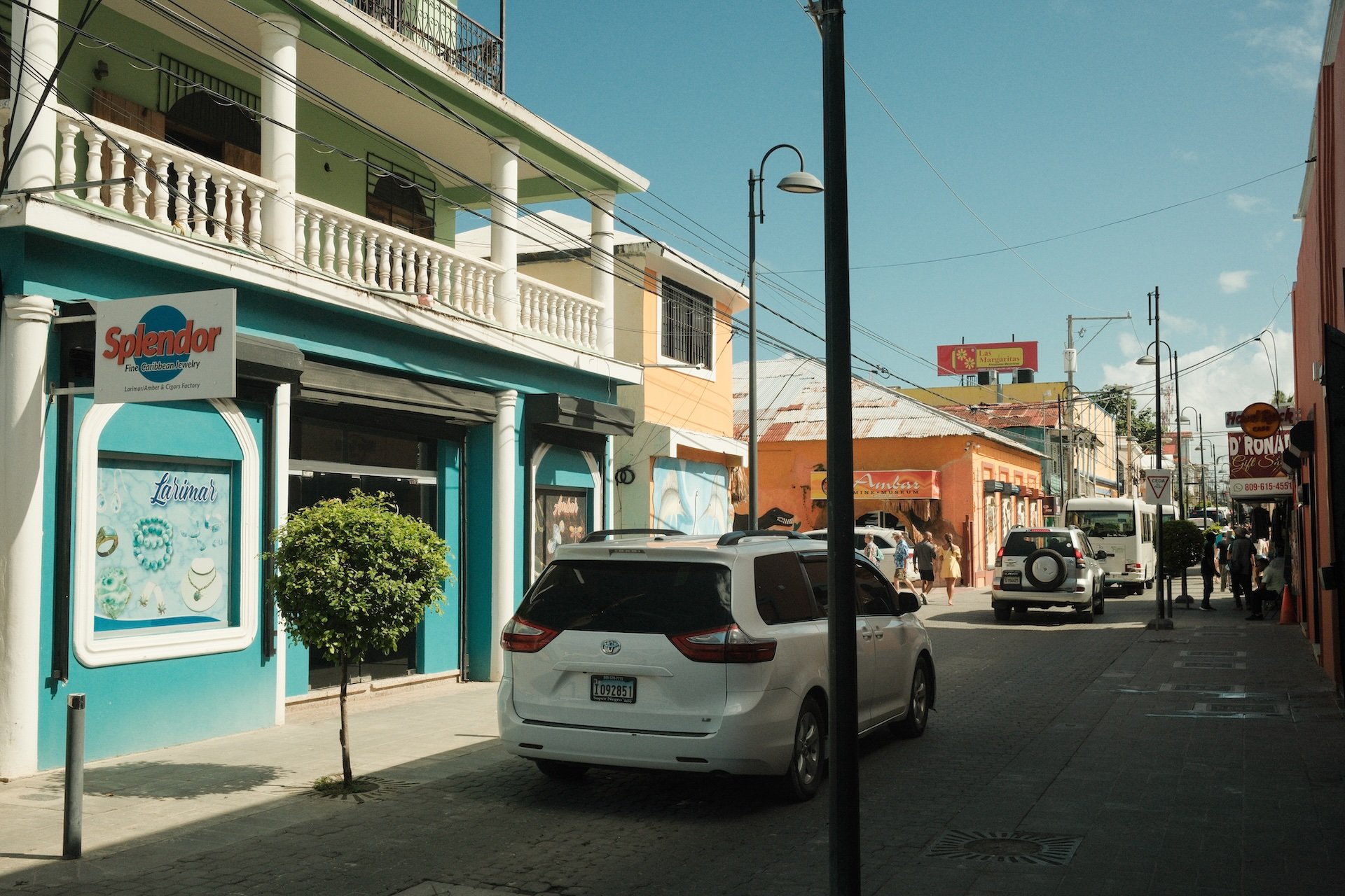 Street scene featuring colorful buildings, parked cars, pedestrians, and various signs on a sunny day.