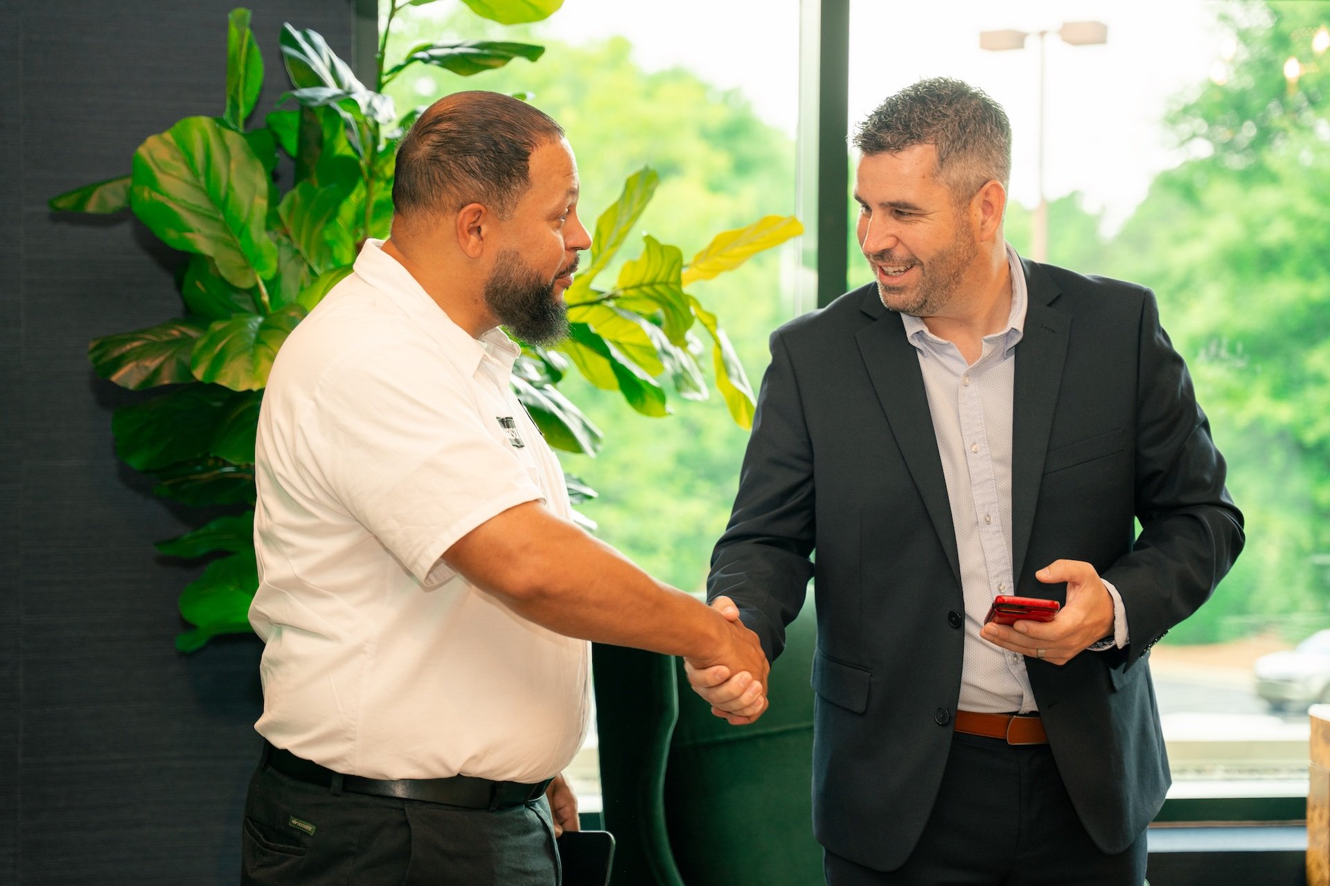 Two men shaking hands in a business setting with a large green plant in the background.