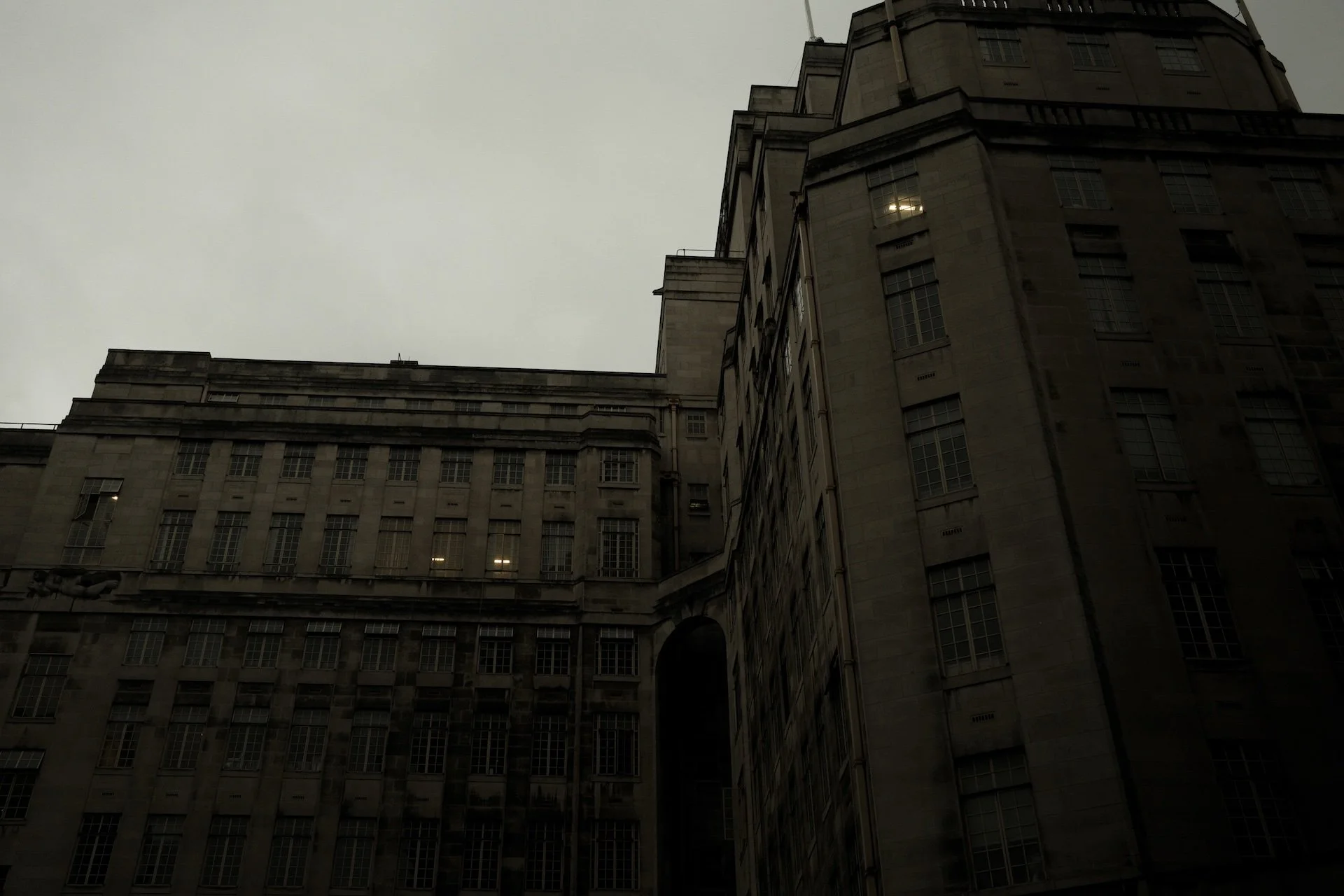 A large, old stone building with many rectangular windows and an archway, seen from a low angle against a cloudy sky.