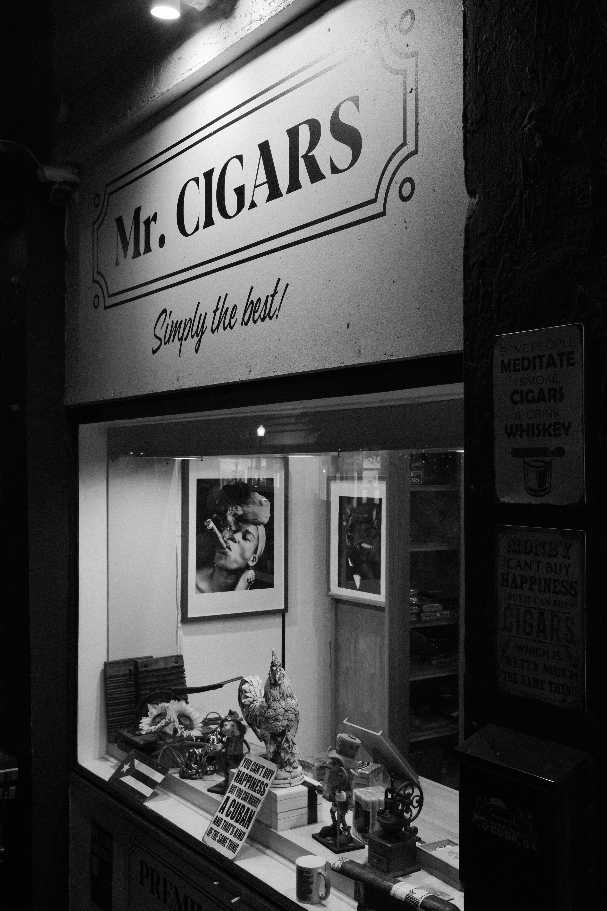 Black and white photo of a storefront window with a sign that reads 'Mr. Cigars' and 'Simply the best!' inside, decorative items and framed pictures, and two signs on the side advertising cigars and whiskey.