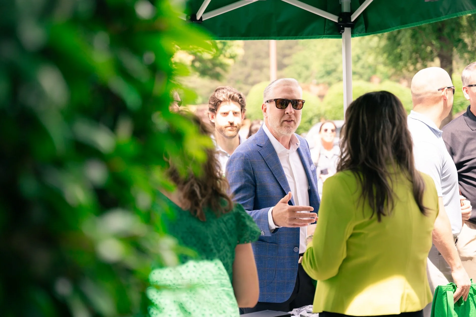 Group of people outdoors under a green canopy, engaging in conversation, some wearing sunglasses and business attire.