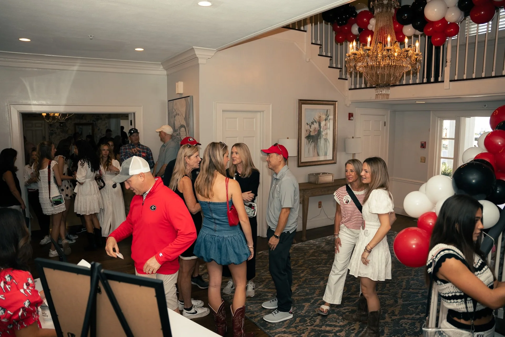 Group of people at a party, some wearing University of Georgia and Cincinnati Bengals gear, decorated with red, white, and black balloons, in a well-lit room with chandelier and artwork on the wall.
