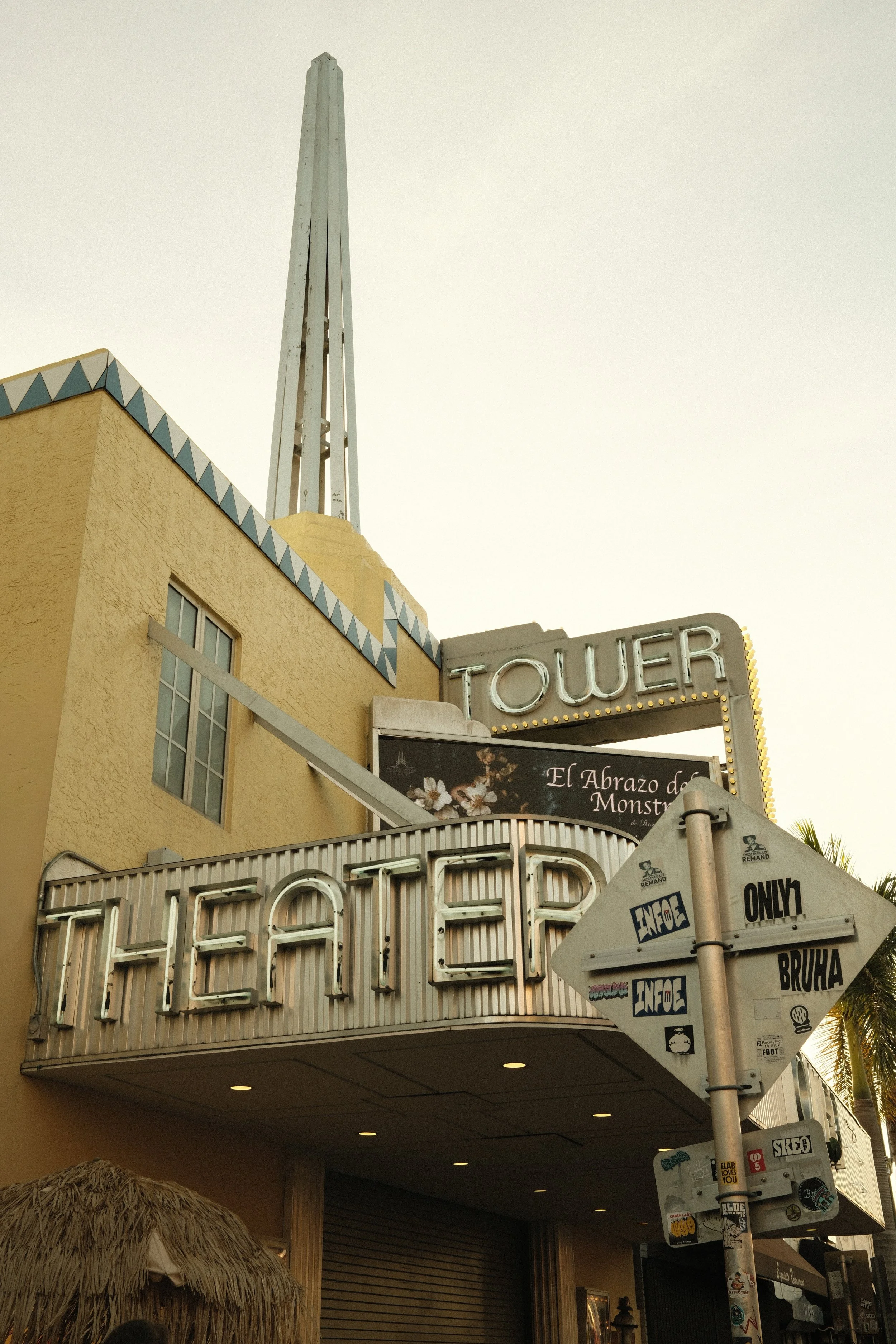 A vintage-style building with a large neon sign reading 'THEATRE' and smaller signs for 'TOWER' and 'EL Abrazo de Monsta.' There is also a smaller arrow sign with various stickers and the words 'ONLY' and 'BRUHA.'