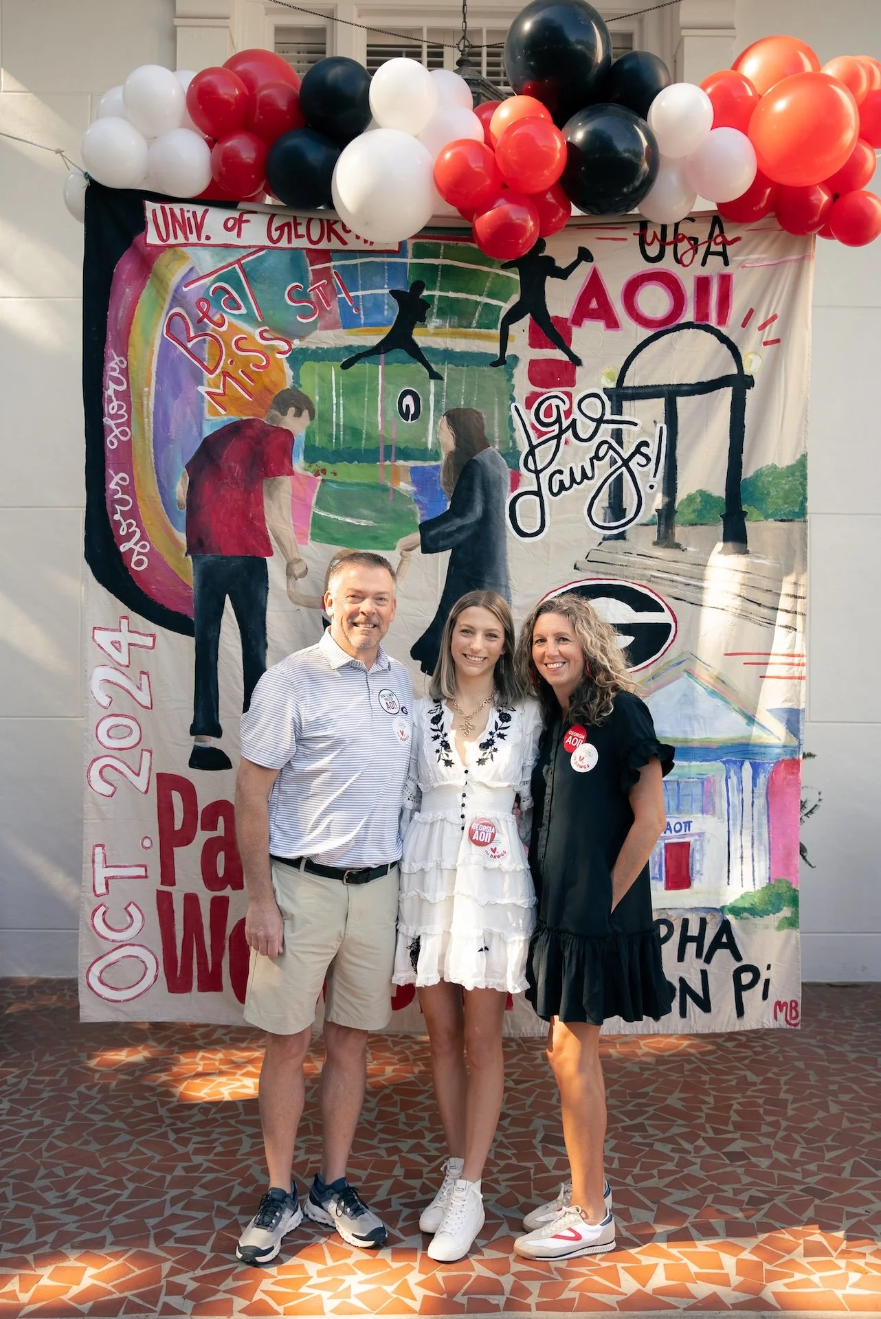 Three people standing in front of a colorful banner decorated with red, white, black, and orange balloons. The banner celebrates an event at the University of Georgia in 2024, with illustrations of people, a rainbow, and graduation-related imagery.