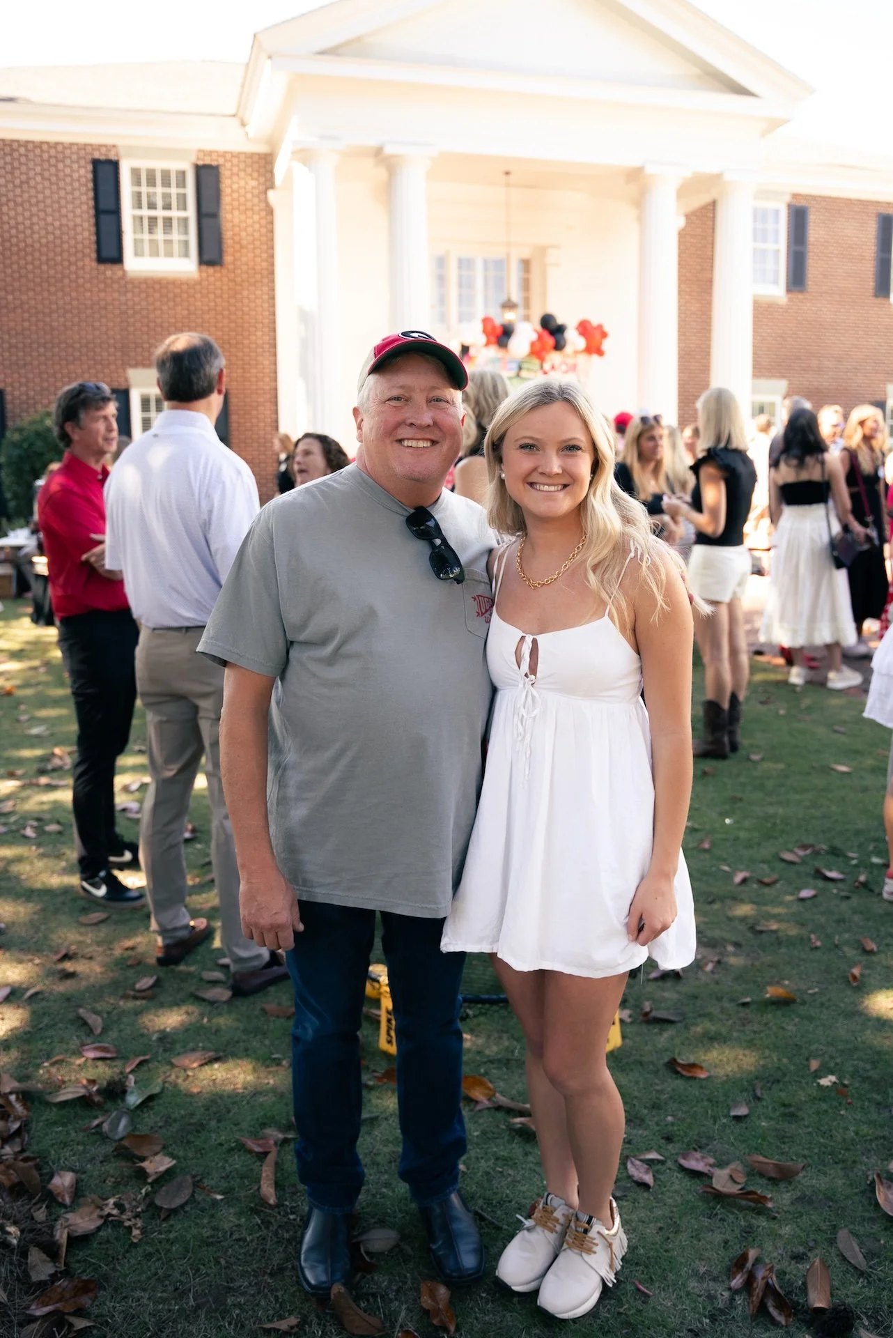 A man and a woman smiling and posing together outdoors at a gathering on a lawn with fallen leaves, with a large house and other people in the background.