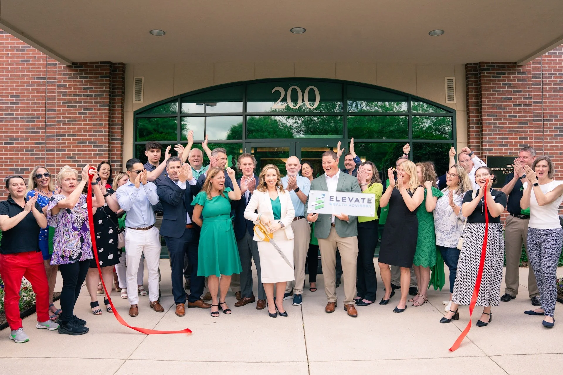 Group of people at a ribbon-cutting ceremony in front of a building numbered 200, celebrating with applause and cheers, holding a sign that reads 'ELEVATE Wealth Advisory'.