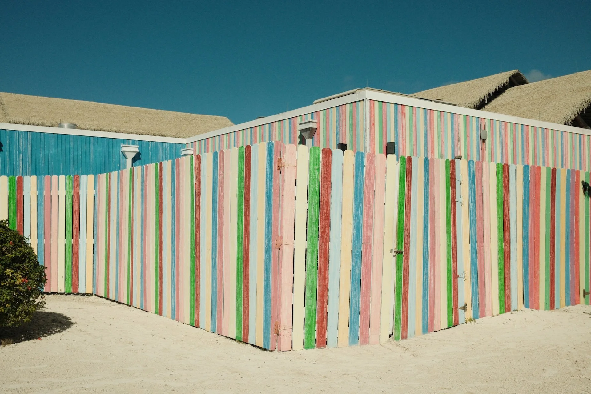 Colorful striped fence on the beach with sand and thatched roof structures in the background.
