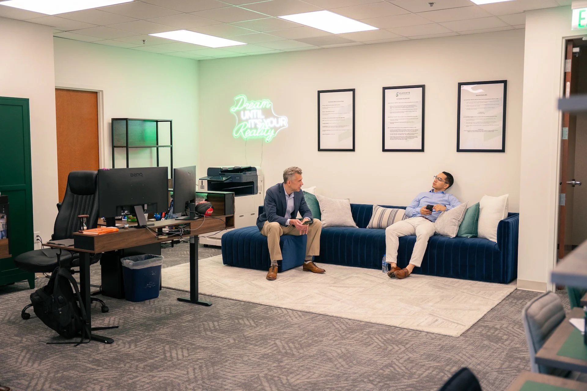 Office lounge with two men having a conversation; one seated on a beige chair, the other on a navy blue velvet sectional; desk with dual monitors, office supplies, and a trash can; wall decor including framed documents and neon sign that reads 'Dream