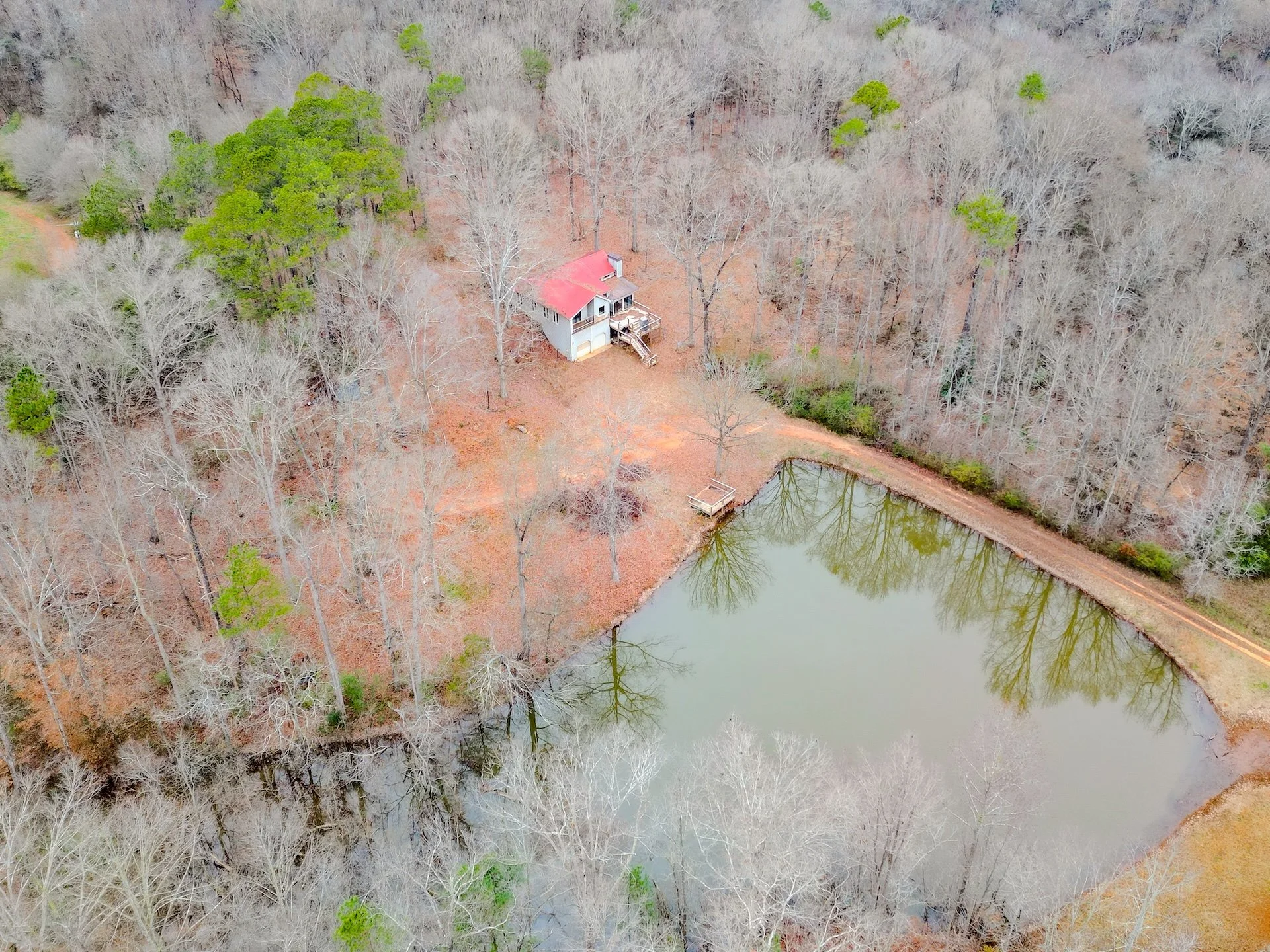 Aerial view of a small house with a red roof situated in a wooded area next to a pond, with trees mostly leafless and a dirt path leading to the house.