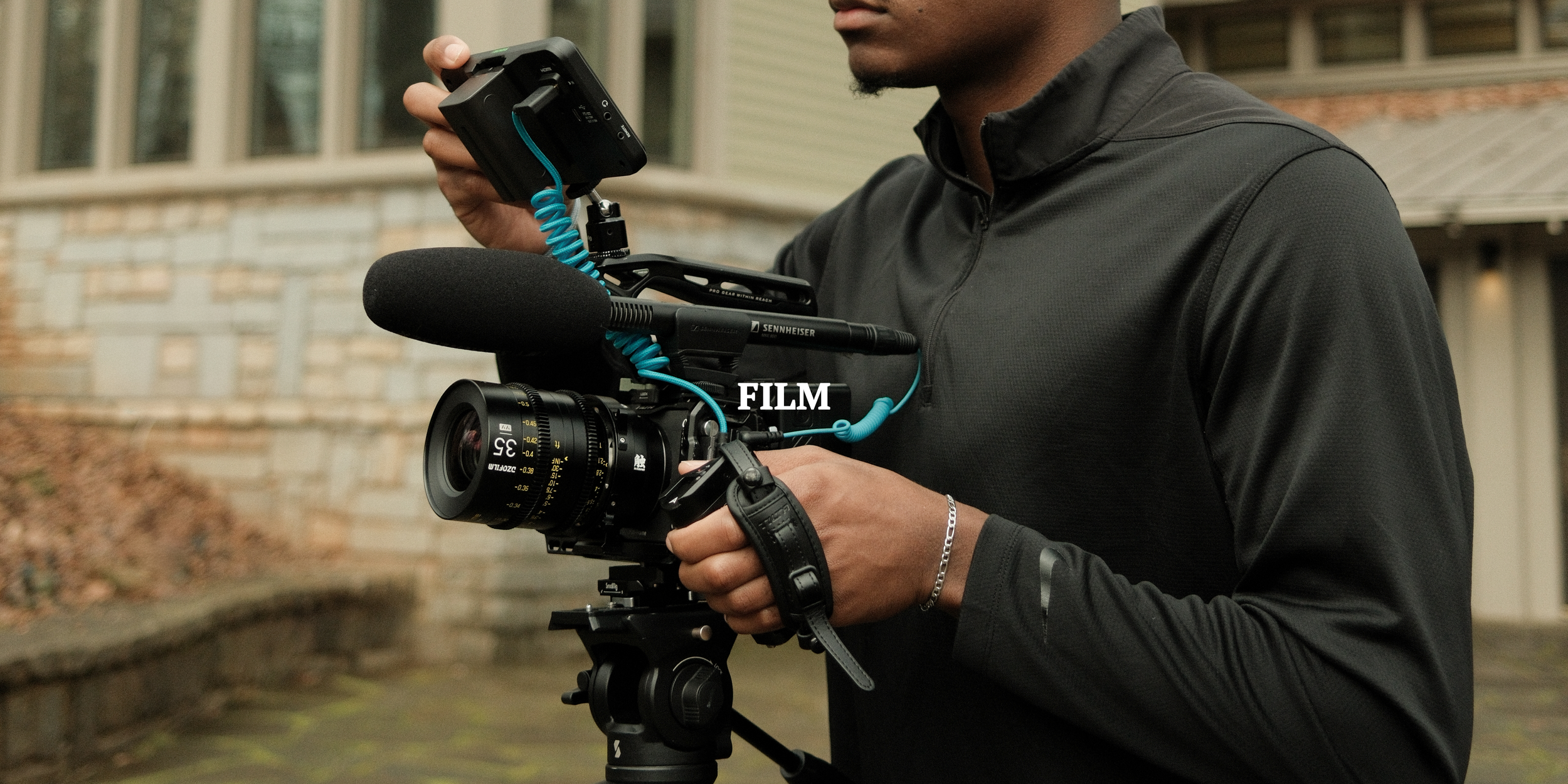 A person holding a professional film camera outdoors, wearing a black jacket, with a building in the background.