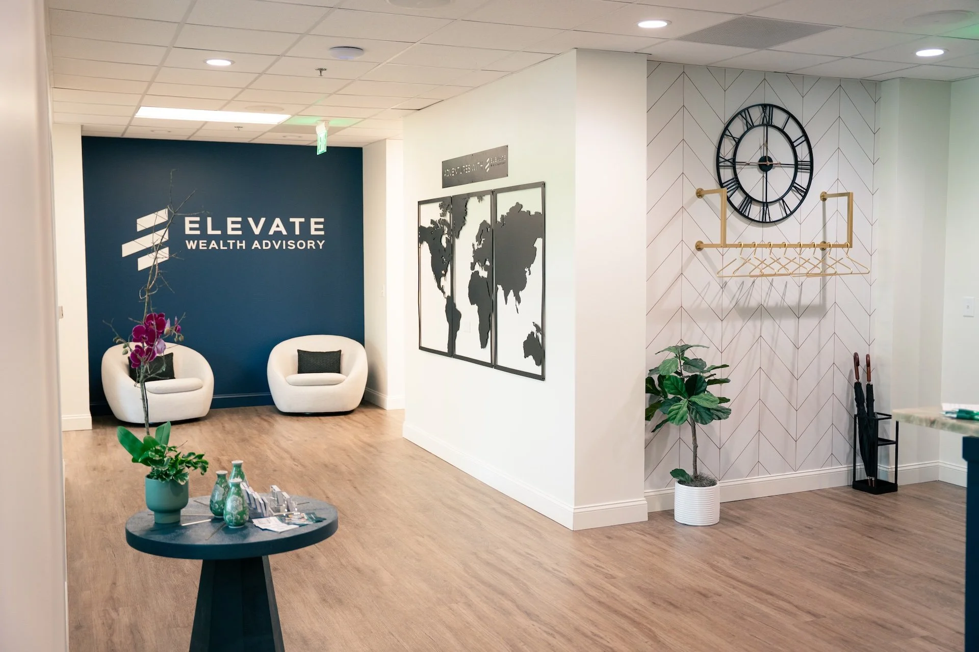 Modern office waiting area with two white chairs, a small round table with flowers and water bottles, a blue accent wall with the name 'Elevate Wealth Advisory', framed world map, decorative clock, potted plants, and umbrella stand.