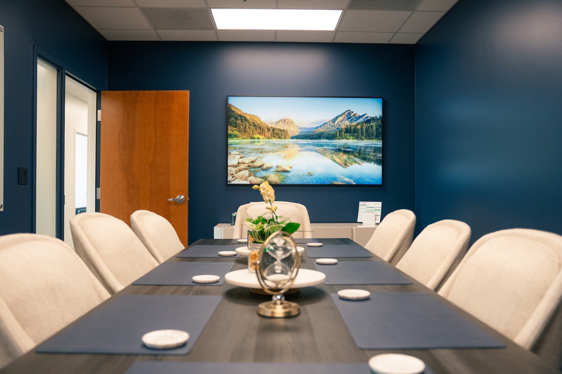 Conference room with a dark wooden table, eight beige chairs, a small centerpiece with a plant and an hourglass, placemats, and a large landscape painting of a river and mountains on a blue wall.