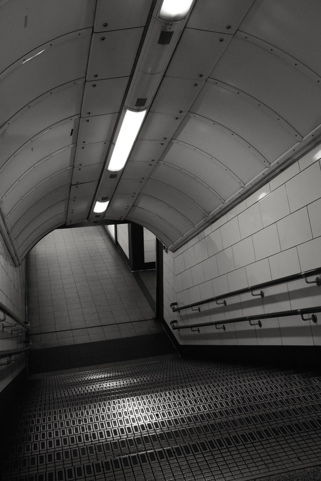 Underground subway station staircase with metal handrails, tiled walls, and ceiling lights.