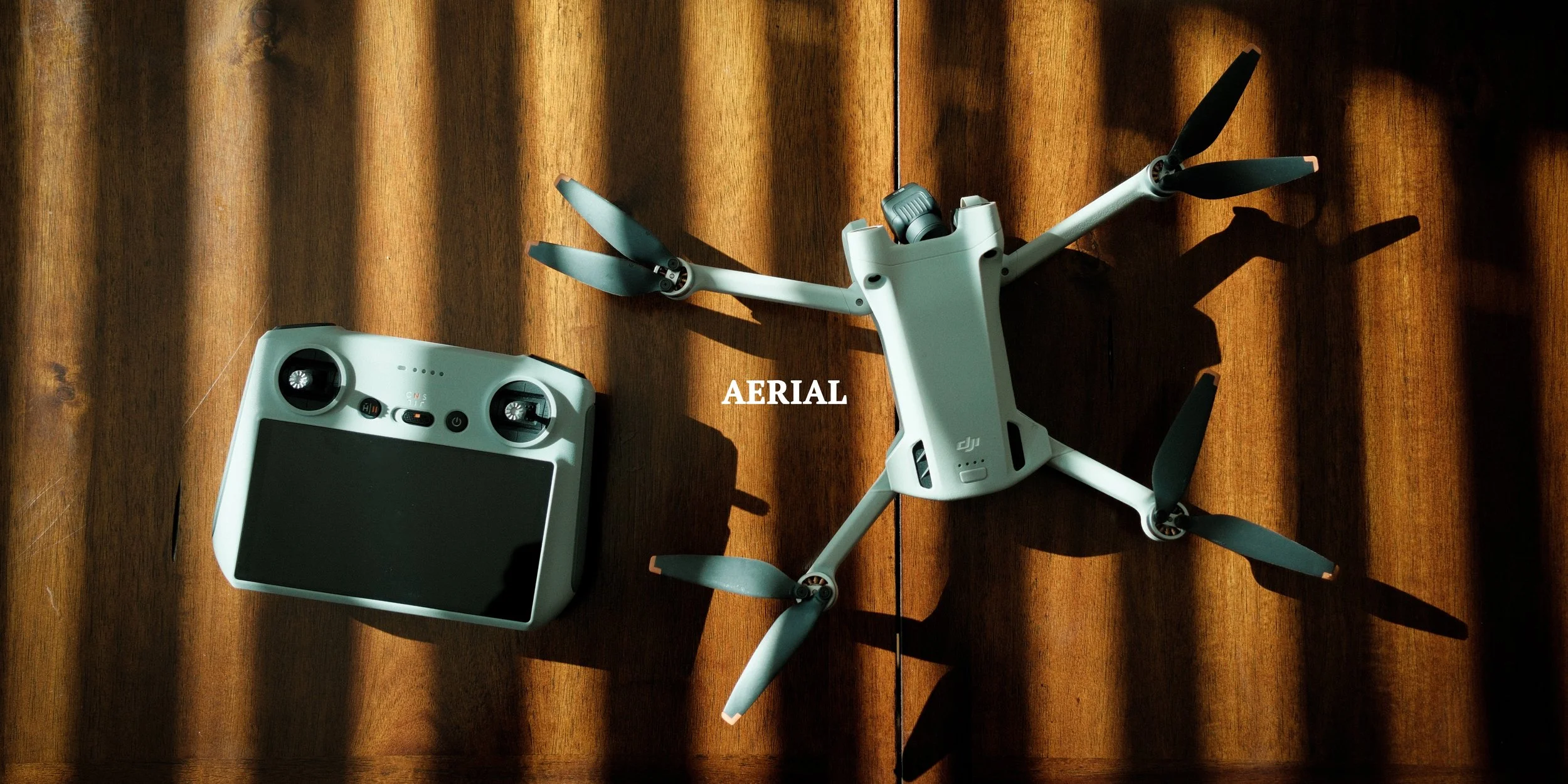 A drone and a remote control on a wooden surface with sunlight casting shadows, with the word 'AERIAL' written in the center.