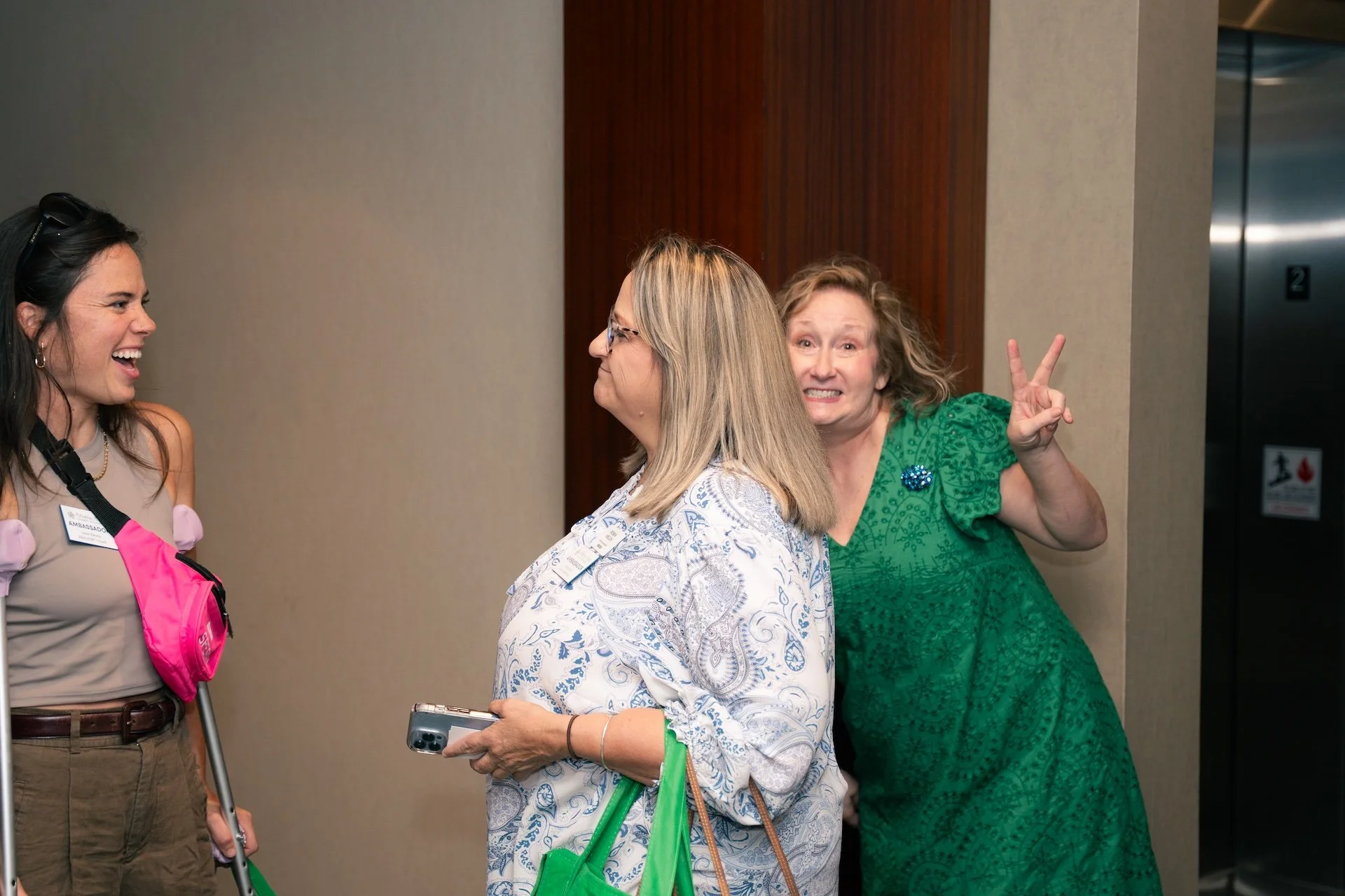Three women interacting and having fun in a hallway. The woman on the right is making a playful expression and holding up a peace sign. The woman in the middle is smiling and holding a phone, while the woman on the left is laughing. They are indoors 