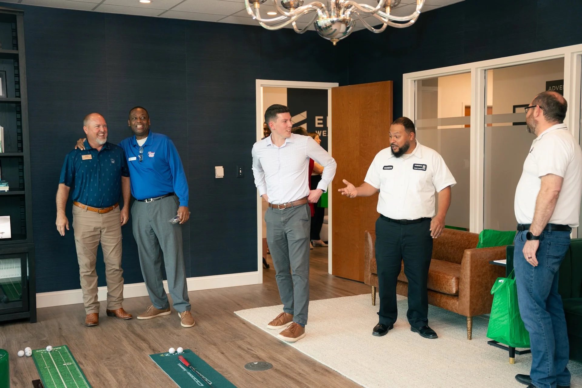 Five men talking and laughing in an office setting, with some golf-related items on the floor, including golf balls, a mini golf putting green, and a golf club.