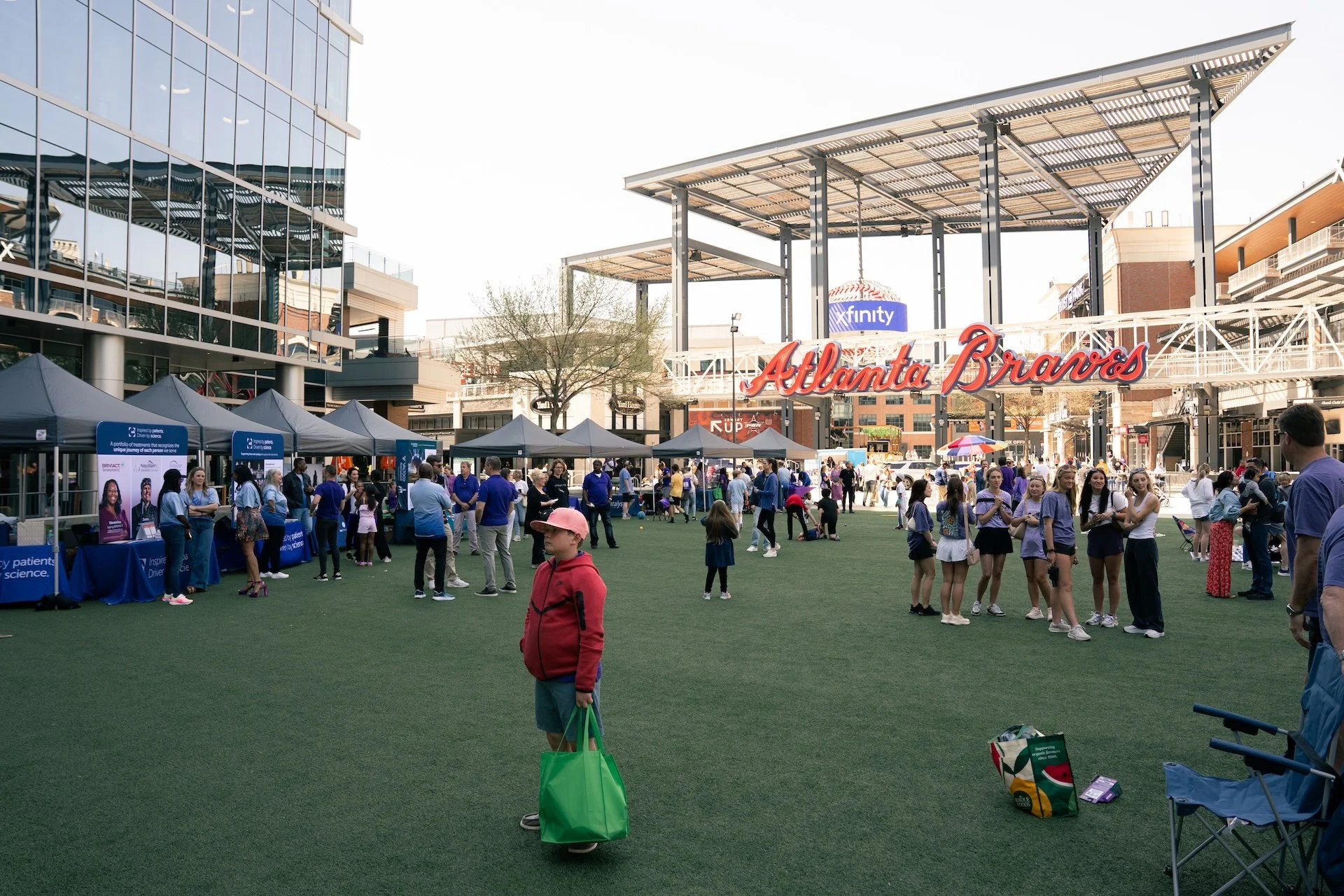 Outdoor scene at Atlanta Braves stadium with people walking, some in group conversations, up for a community event or fair, various booths set up under canopies, large sign reading 'Atlanta Braves' in the background, modern buildings, and a partly cl
