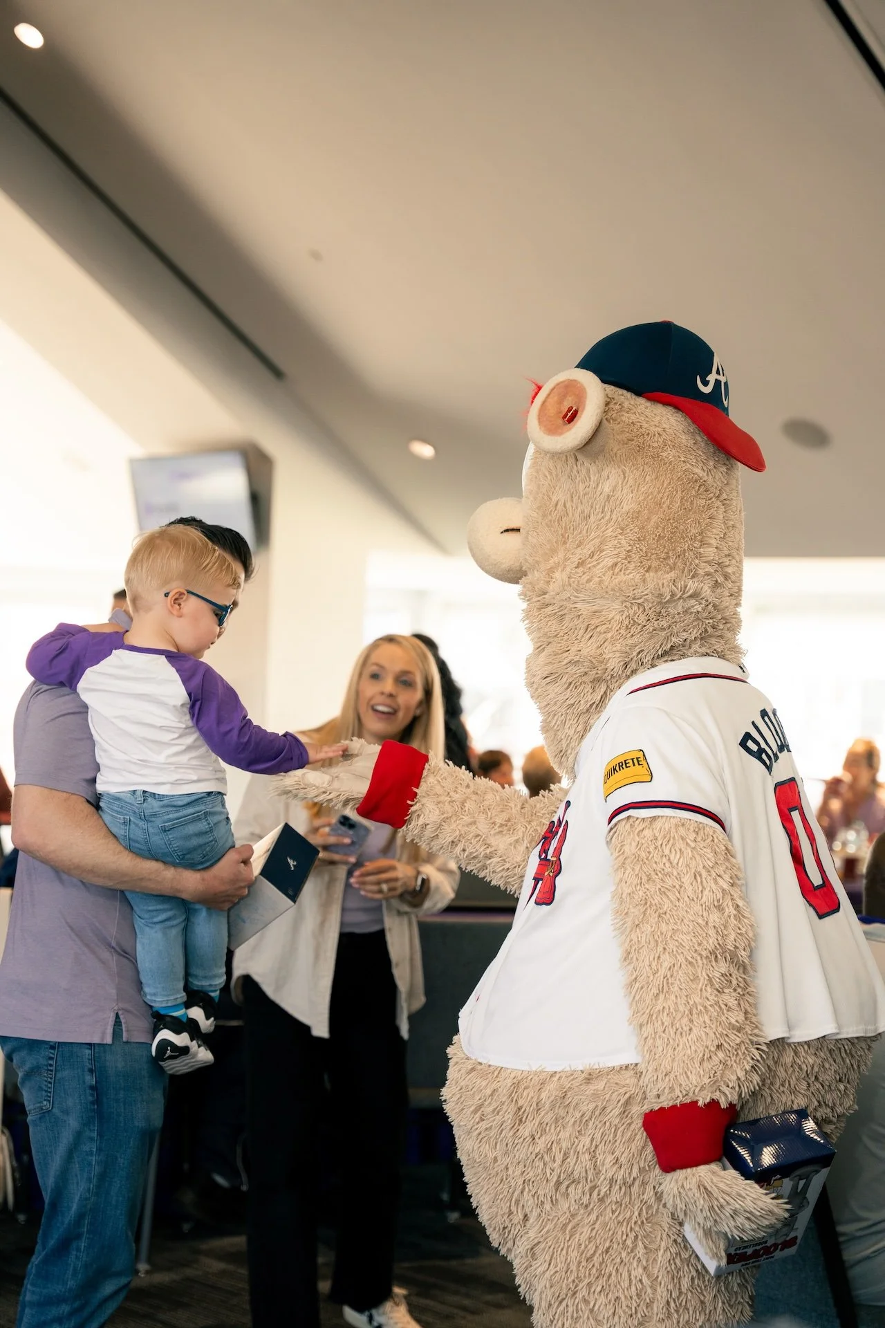 A person in a baseball mascot costume shaking hands with a young boy, while a woman smiles in the background at an indoor event.