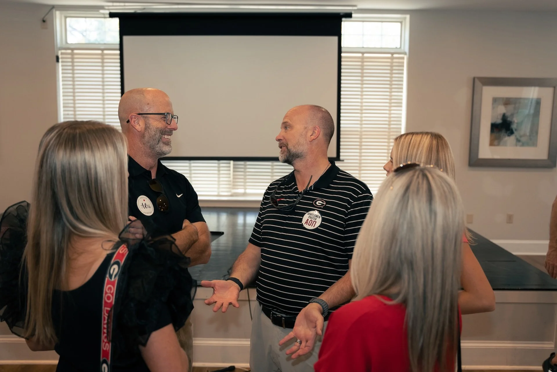 Group of five adults engaging in conversation indoors, with a blank presentation screen and window blinds in the background.