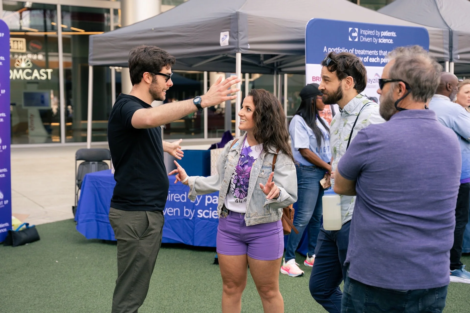 People engaging in conversation at an outdoor event with informational tents and banners.
