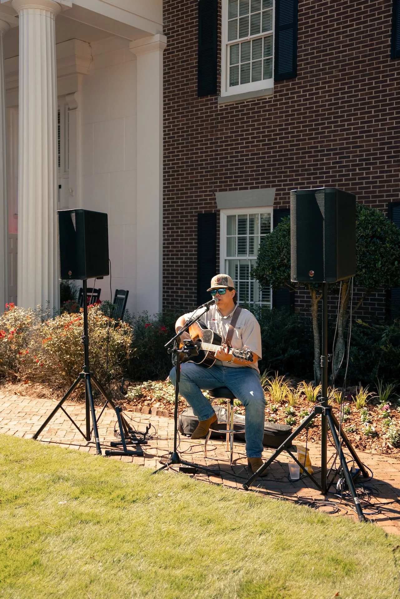 A young man playing an acoustic guitar and singing into a microphone outdoors, with two large speakers on stands beside him, in front of a brick house with white columns and windows.