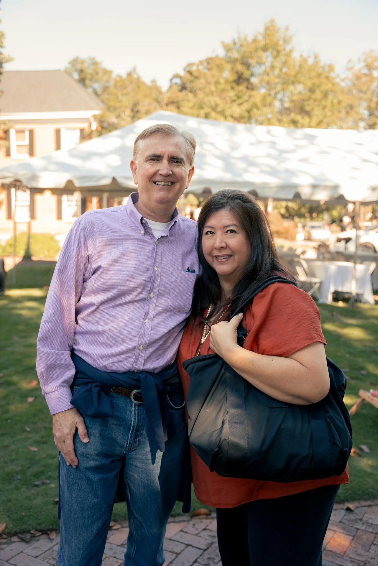 A man and woman standing outdoors during daytime, smiling, with a white canopy tent and trees in the background.