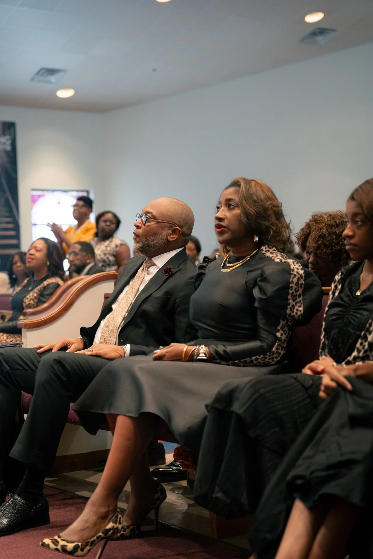 People seated in an audience at a formal event, with two individuals in the foreground. The man is wearing a dark suit, light-colored shirt, and patterned tie, and the woman is dressed in a black dress with animal print accents and leopard print high