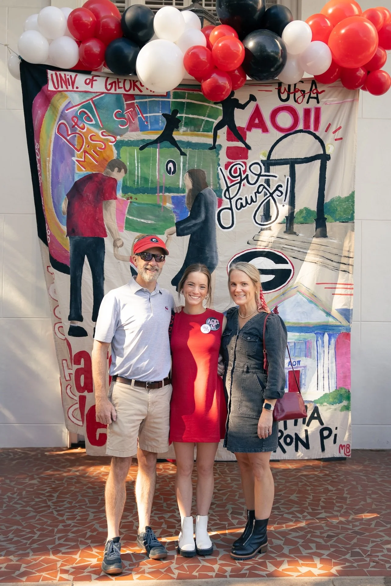 Three people standing in front of a colorful banner with balloons on top. The banner features illustrations and text related to the University of Georgia and their athletic programs, including images of athletes and a football stadium.